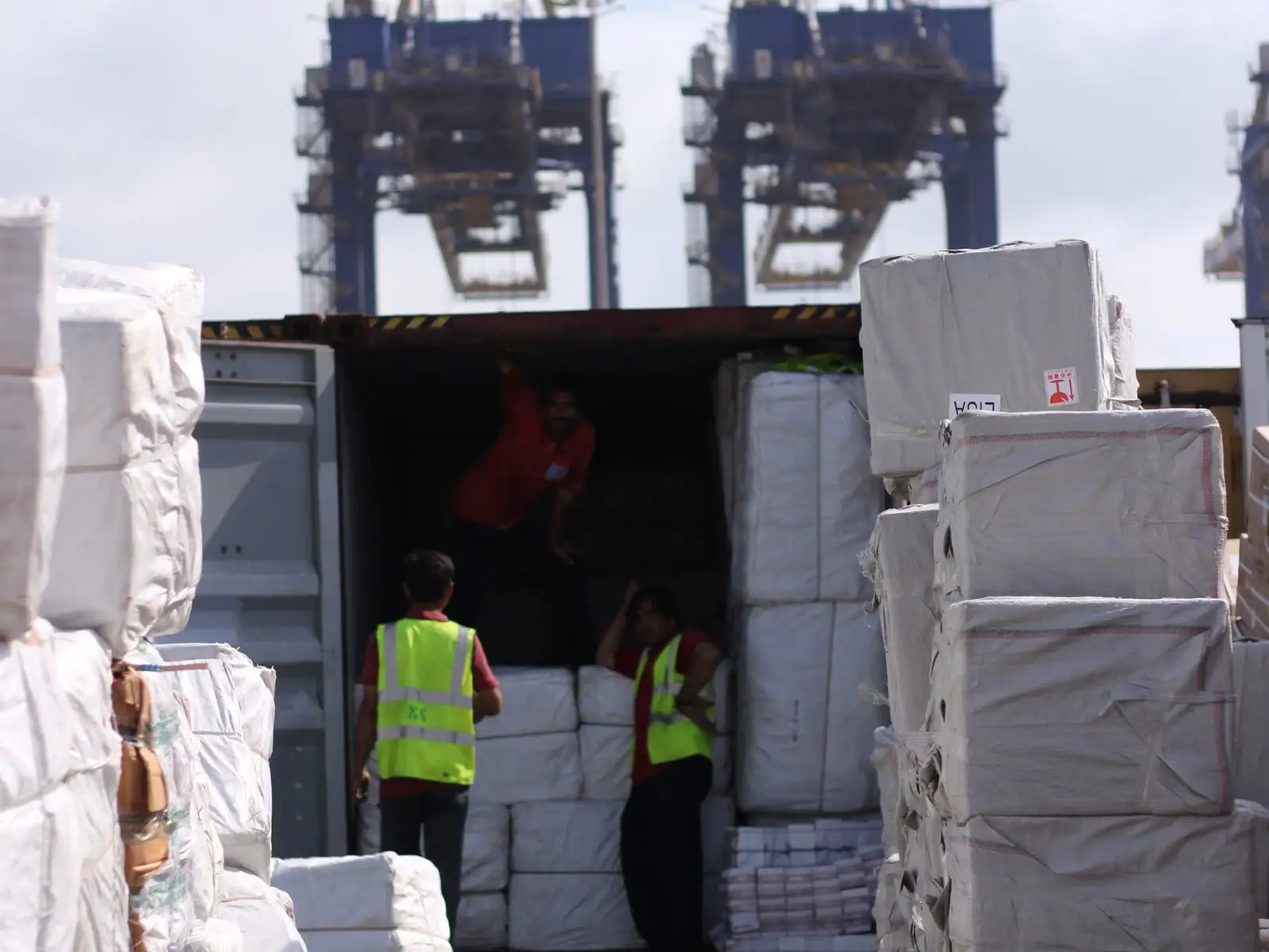 Bales of woven bags being loaded into a shipping container at port, gantry cranes behind